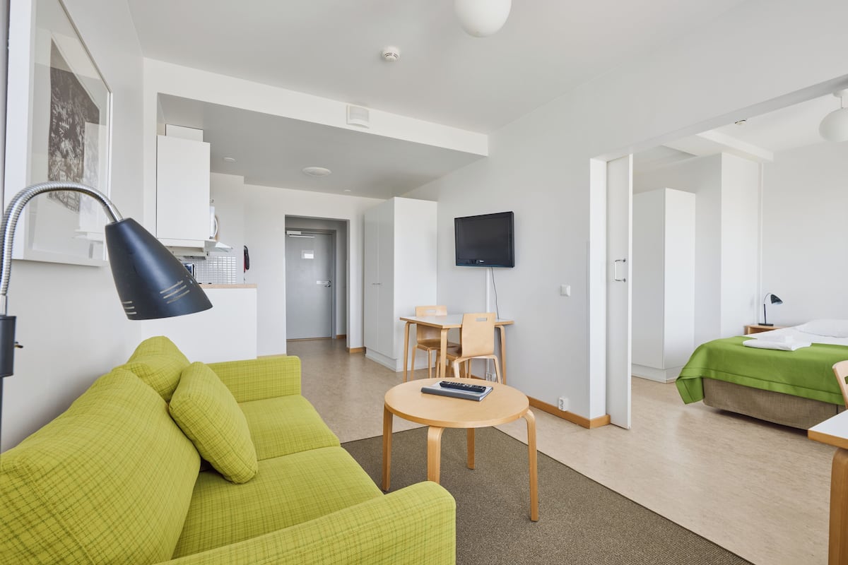 A modern hotel room with a green sofa, a round wooden table, a flat-screen TV, and a bed with a green bedspread.