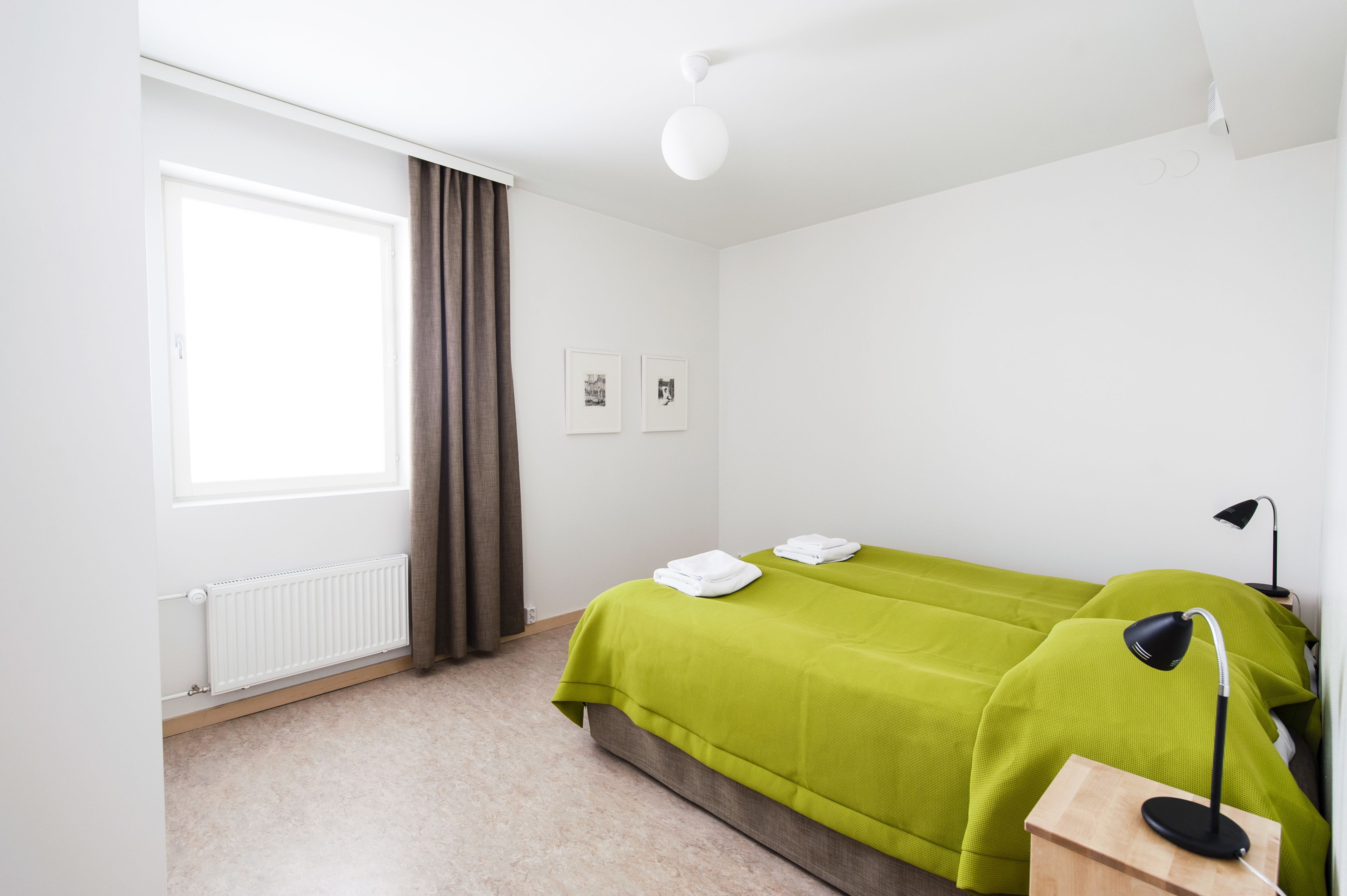 A hotel room with a bed, a nightstand, a lamp, a window with curtains, and a radiator.