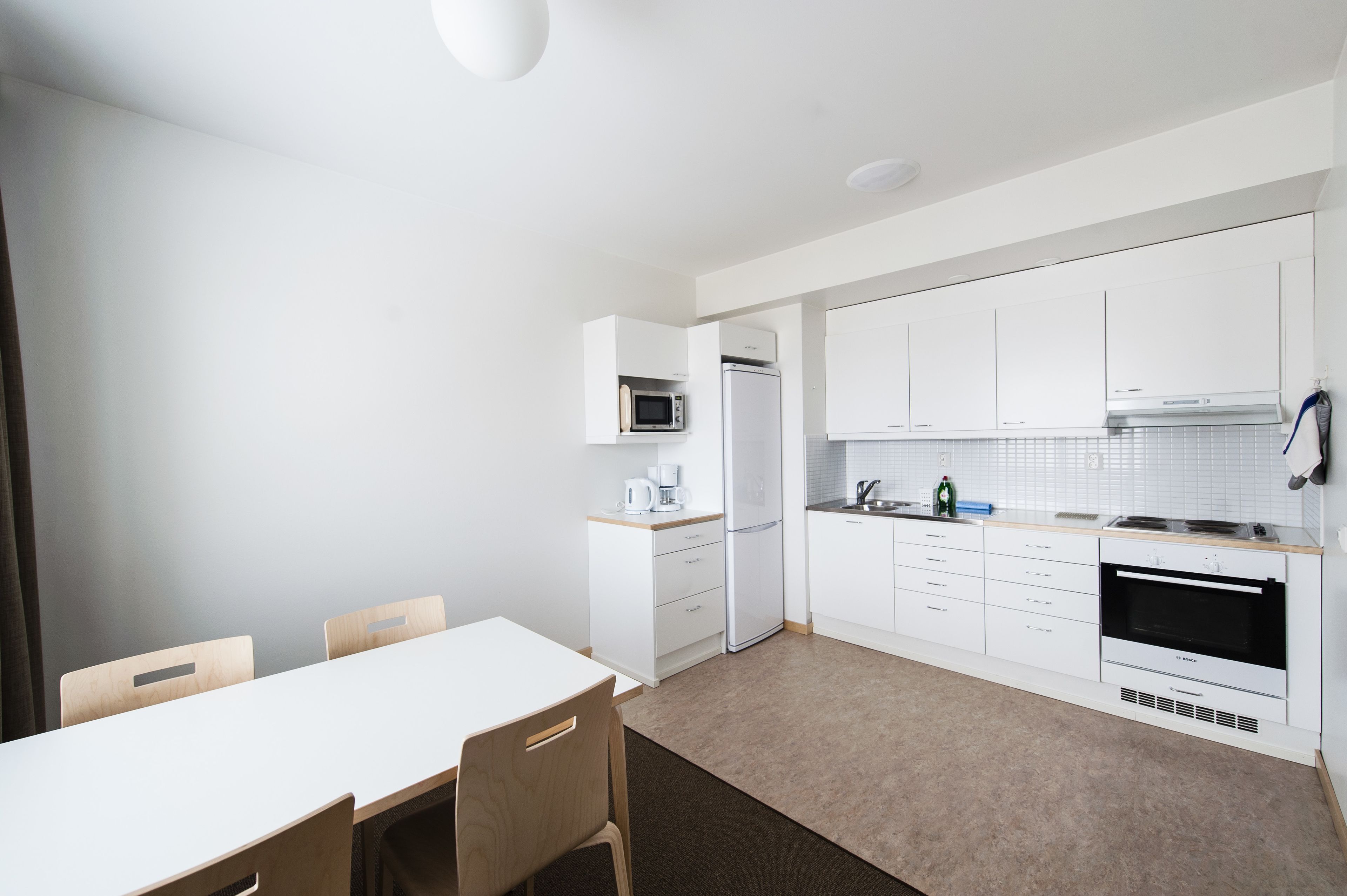 A modern kitchen with white cabinets, a microwave, a coffee maker, and a built-in oven. There is a dining area with a table and chairs.