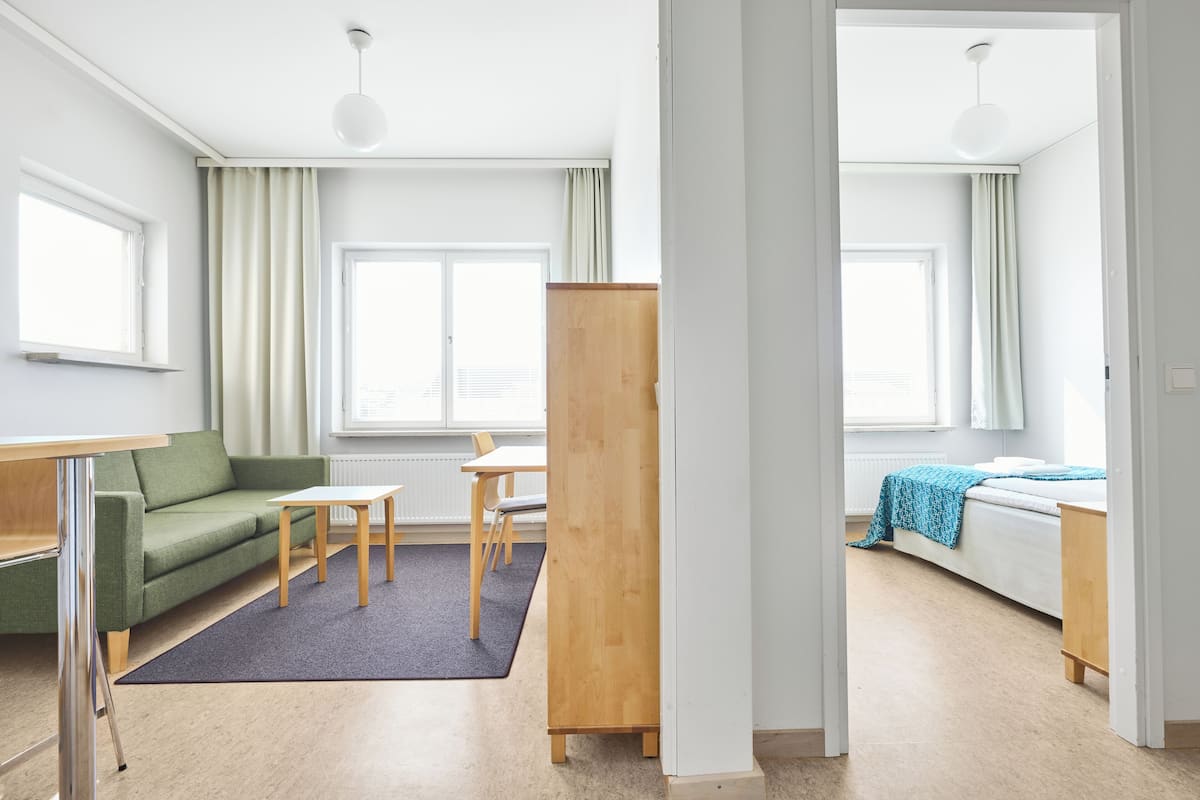 A modern, well-lit room with a green sofa, a wooden table, a small desk, and a bed with a blue blanket.