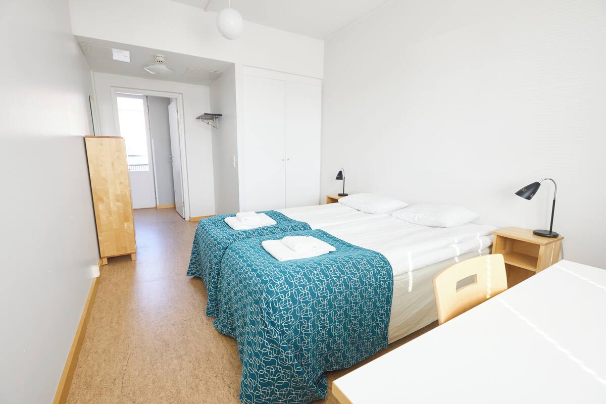A room with two beds, a desk, and a chair. There is a wardrobe and a door leading to another room.