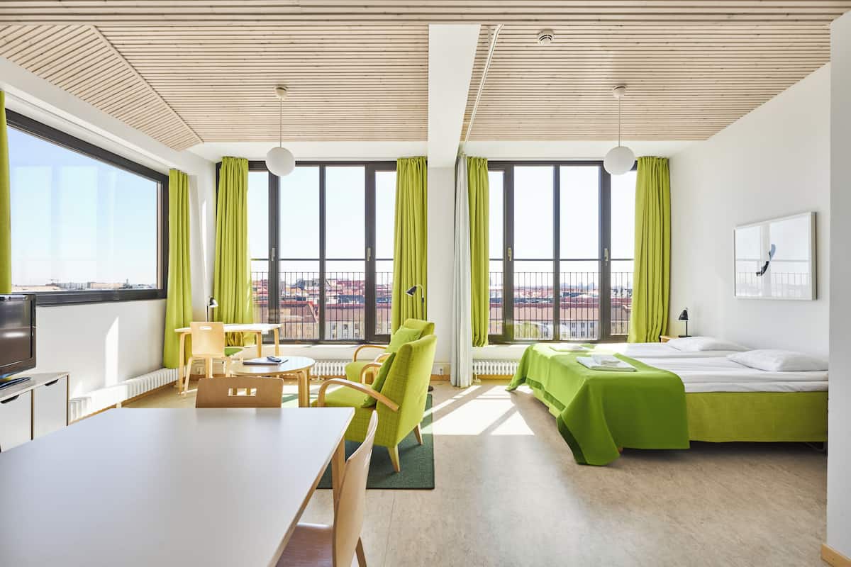 A modern hotel room with a large window, a desk, a chair, a bed, and a small table.