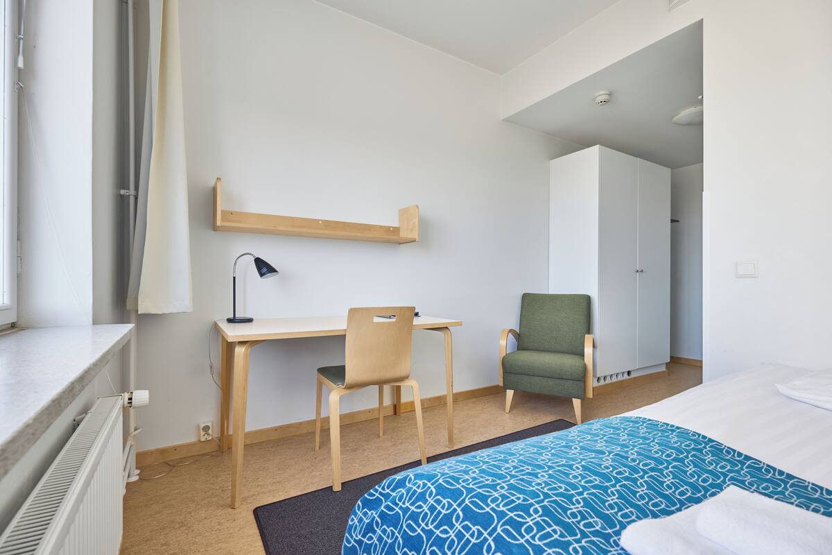 A hotel room with a bed, a desk, a chair, a wardrobe, and a radiator.