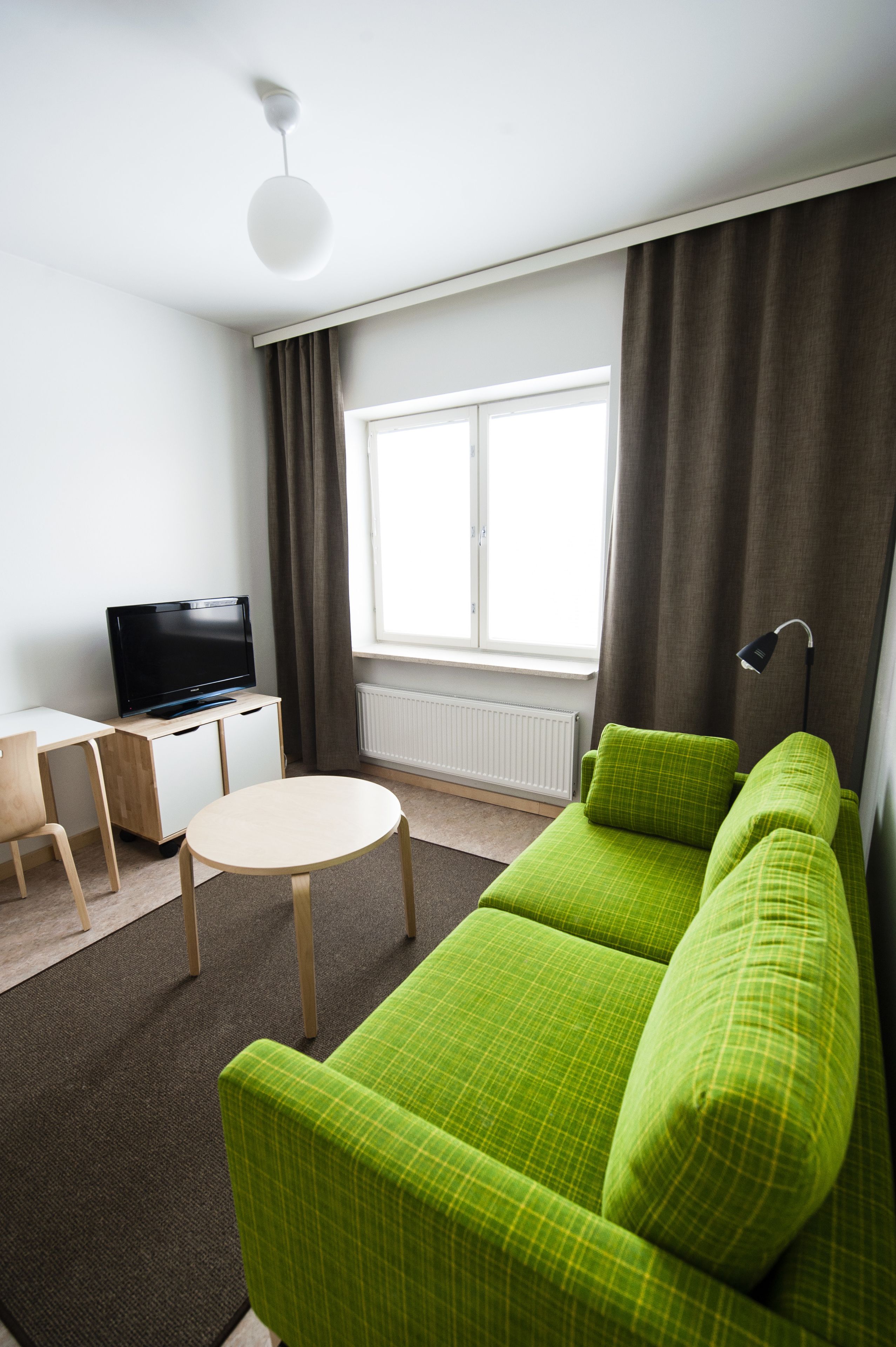 Un salon moderne avec un canapé vert, un téléviseur posé sur un meuble en bois, une table ronde et une grande fenêtre avec des rideaux.