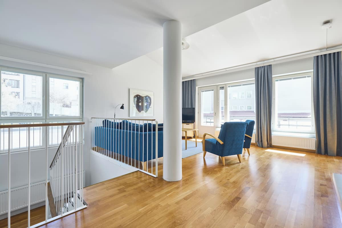 A spacious living area with a wooden floor, a blue sofa, a blue armchair, and a large window with curtains.