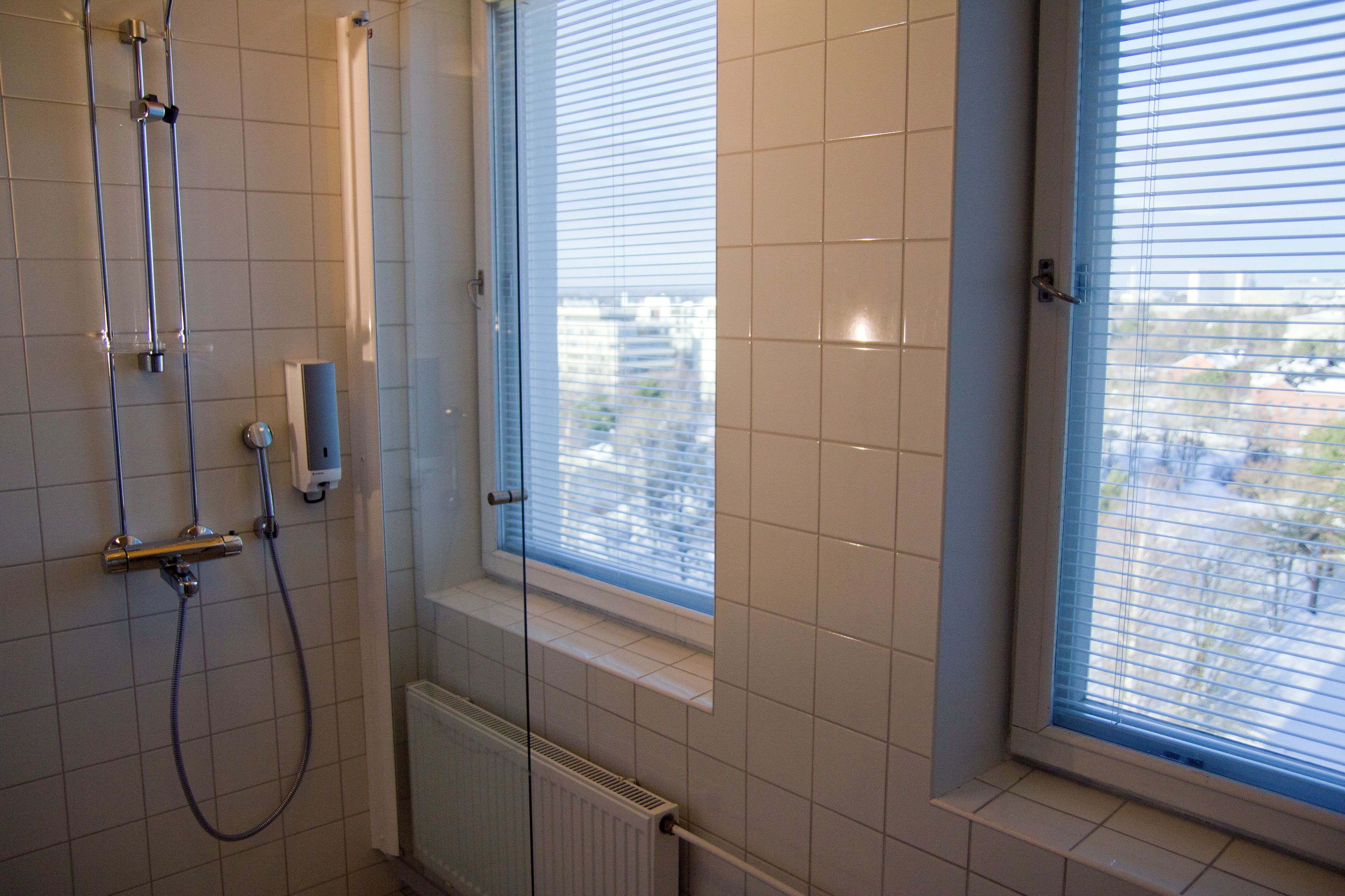 A bathroom with a shower, a window with blinds, and a view of buildings outside.