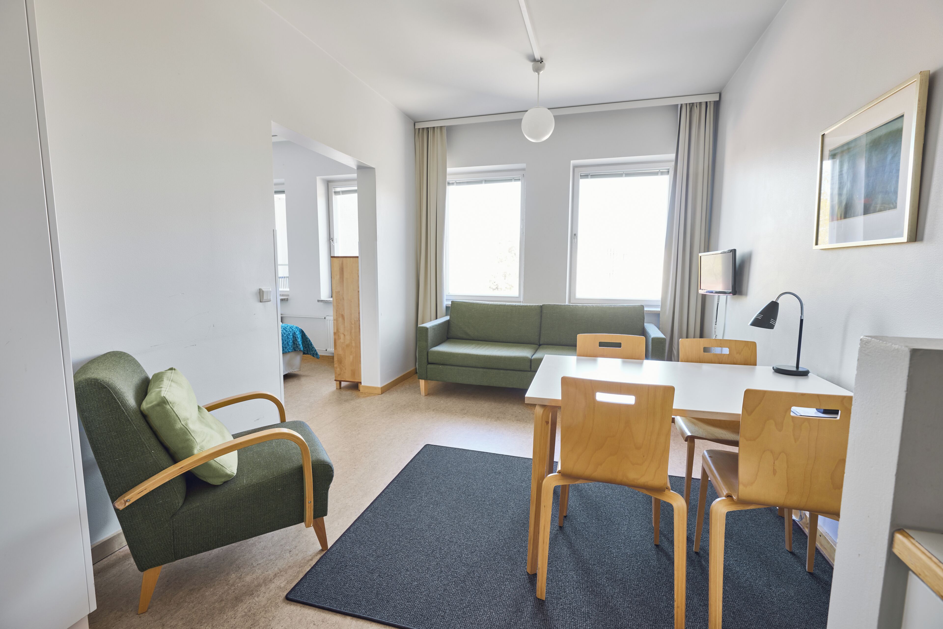 A modern, well-lit room with a green sofa, wooden chairs, a small table, and a TV on the wall.