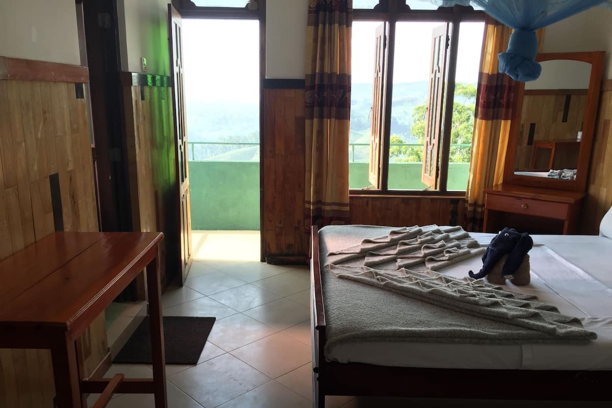 A hotel room with a bed, a wooden desk, and a view of greenery through the window.