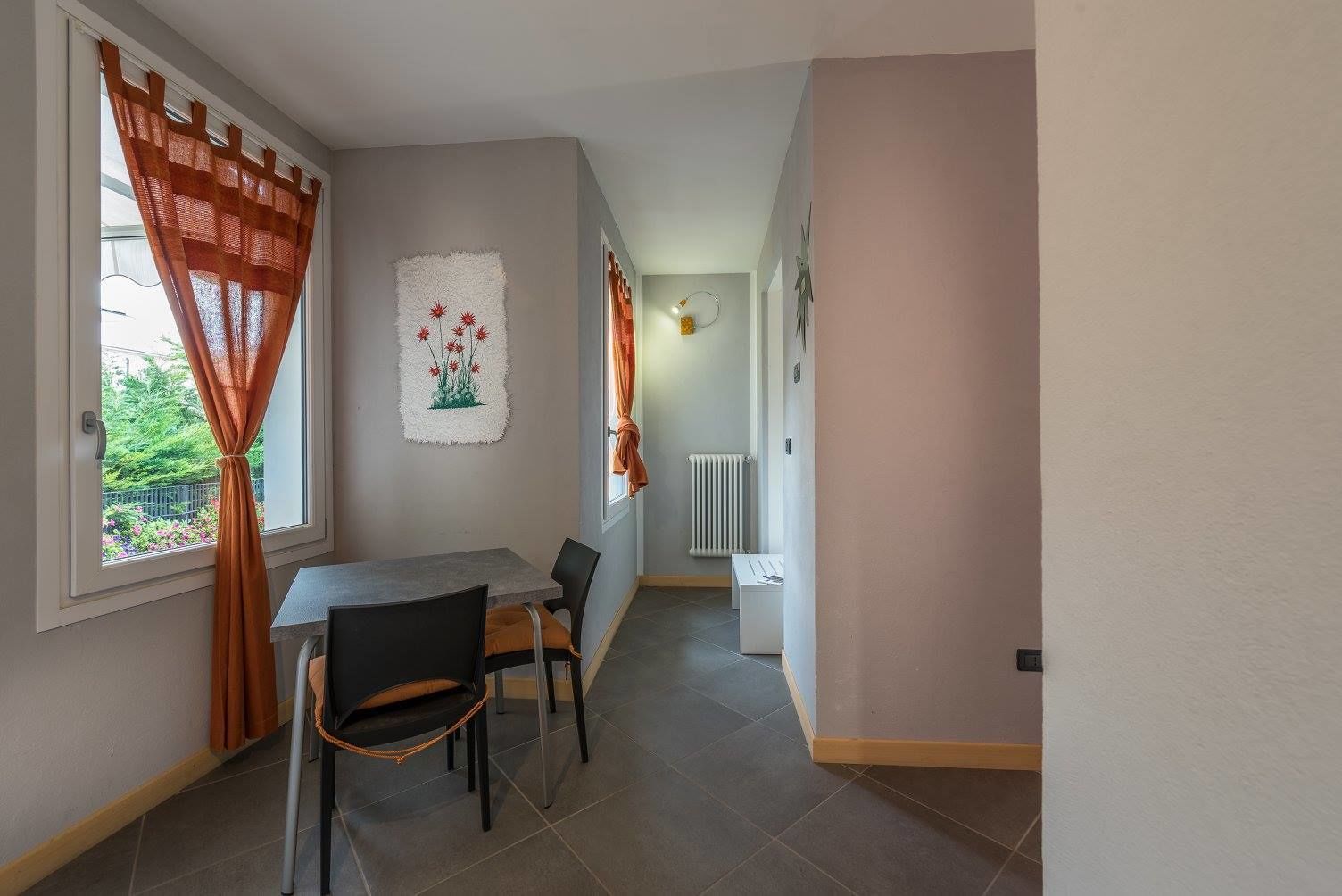Un coin repas avec une table et des chaises, une fenêtre avec des rideaux orange, un radiateur et une œuvre d’art accrochée au mur.