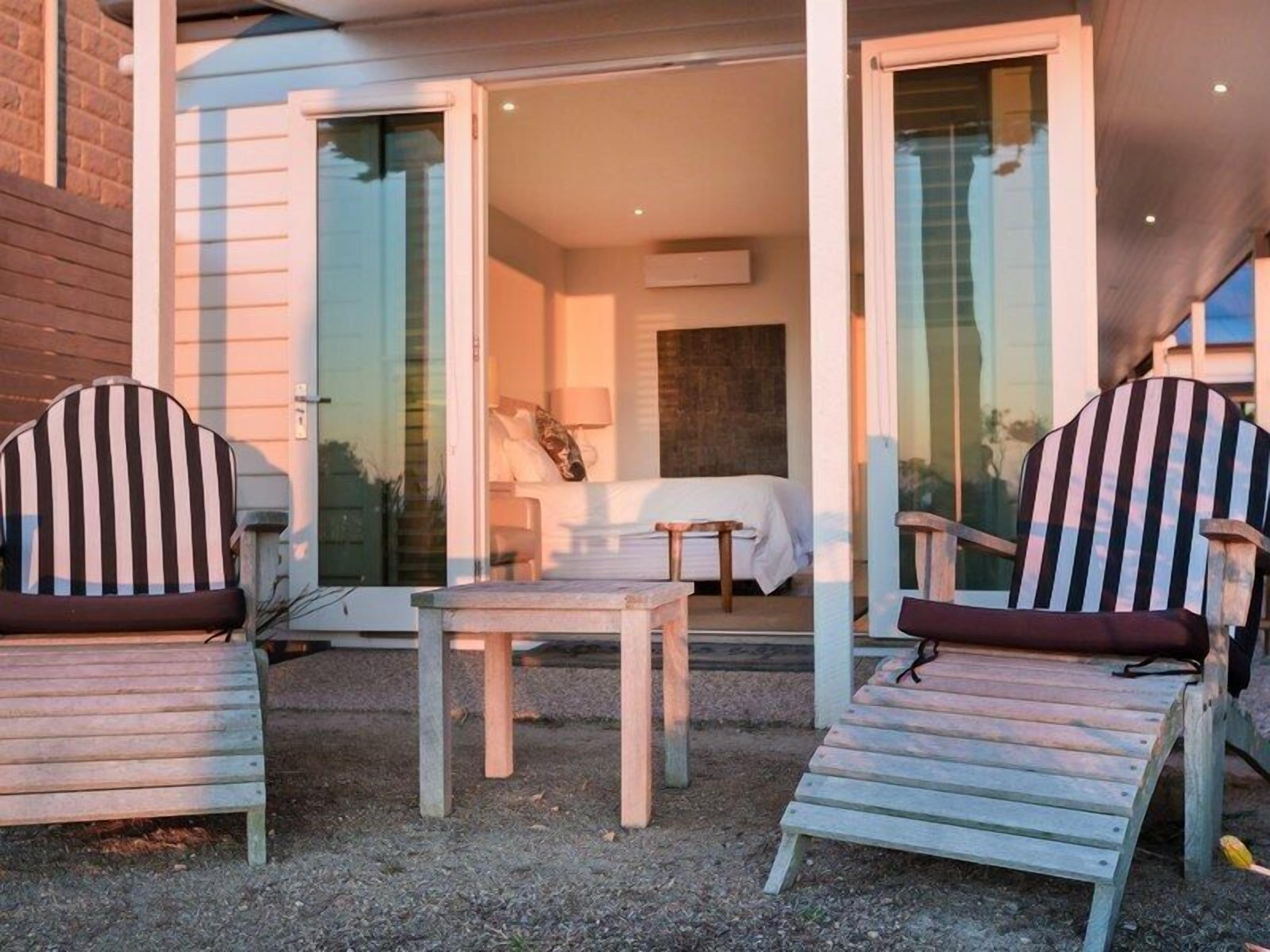 A cozy outdoor seating area with two striped chairs and a small table, leading to a glass door entrance to a bedroom with a bed and a nightstand.