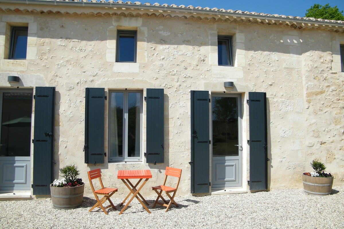 A building with a stone facade, two windows with shutters, and a small table with chairs in front.