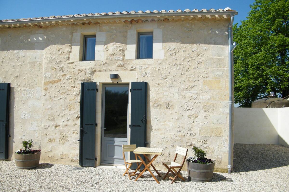 A rustic stone building with a tiled roof, a wooden door, and a small balcony. There are two potted plants and a wooden table with chairs outside.