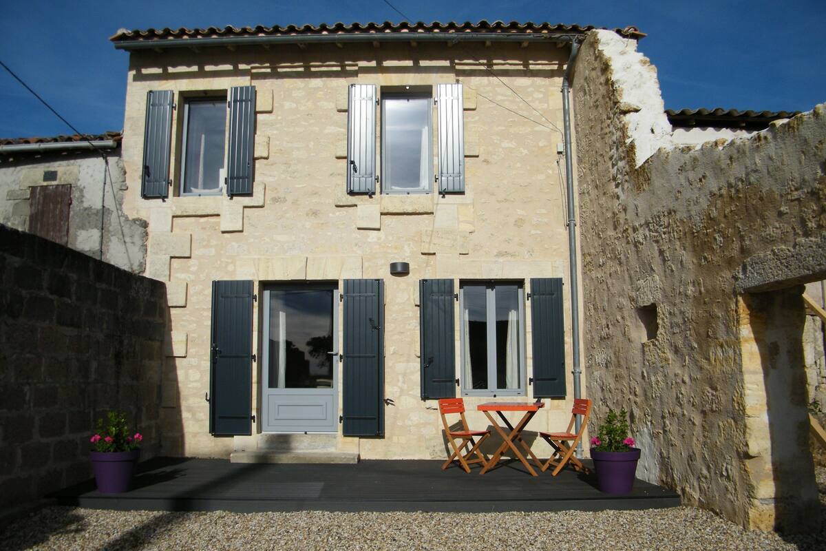 A two-story stone building with shutters, a door, and a small patio with two chairs and two potted plants.