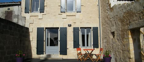 A two-story stone building with shutters, a door, and a small patio with two chairs and two potted plants.