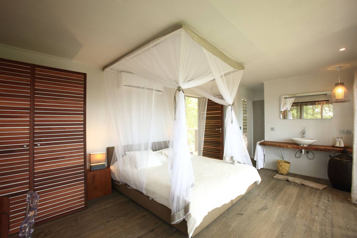Un dormitorio con cama a dosel, suelo de madera, una mesita de noche con lámpara, un espejo grande sobre el lavabo y un armario de madera.