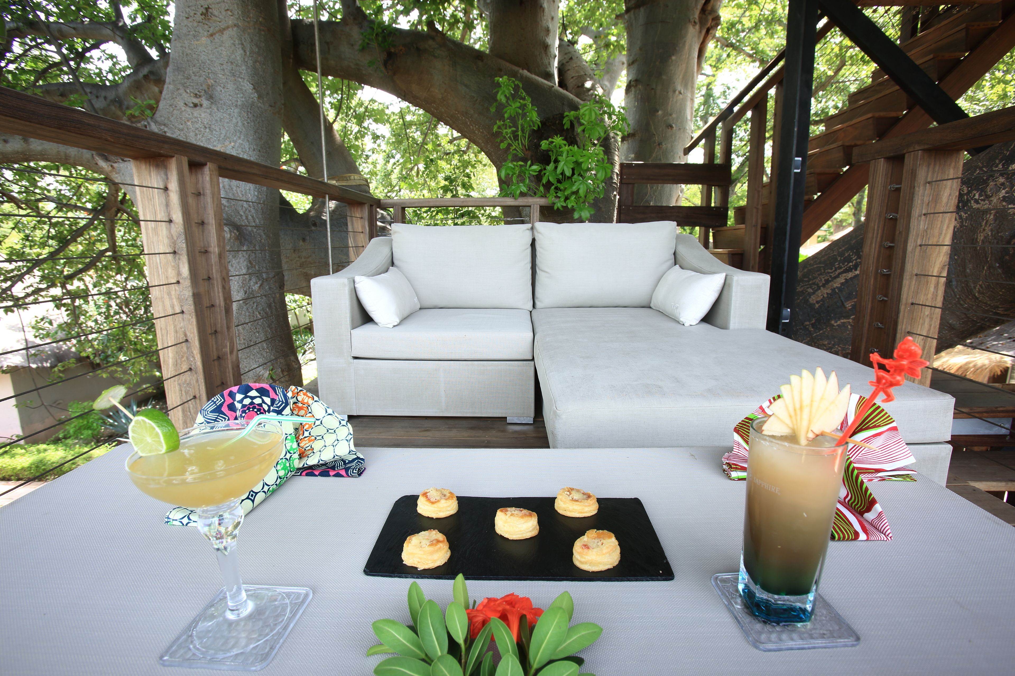 Un área de estar al aire libre con un sofá blanco, una mesa con bebidas y aperitivos, y una estructura de pérgola de madera.