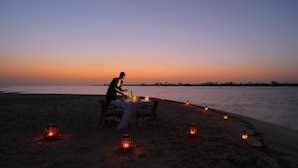 Mesa en la playa con una persona cenando, rodeada de faroles, bajo un cielo al atardecer.