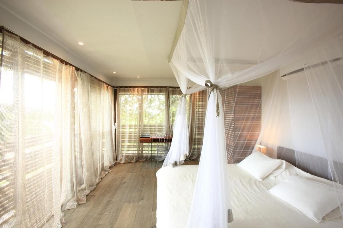 A bedroom with a canopy bed, large windows with sheer curtains, and a wooden floor.