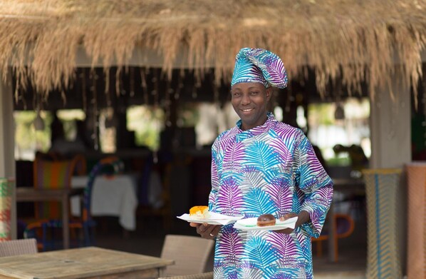 A person in traditional attire holding a plate with food, standing in an outdoor setting with thatched roofing and furniture. - Hotel Les Paletuviers (Fatick)