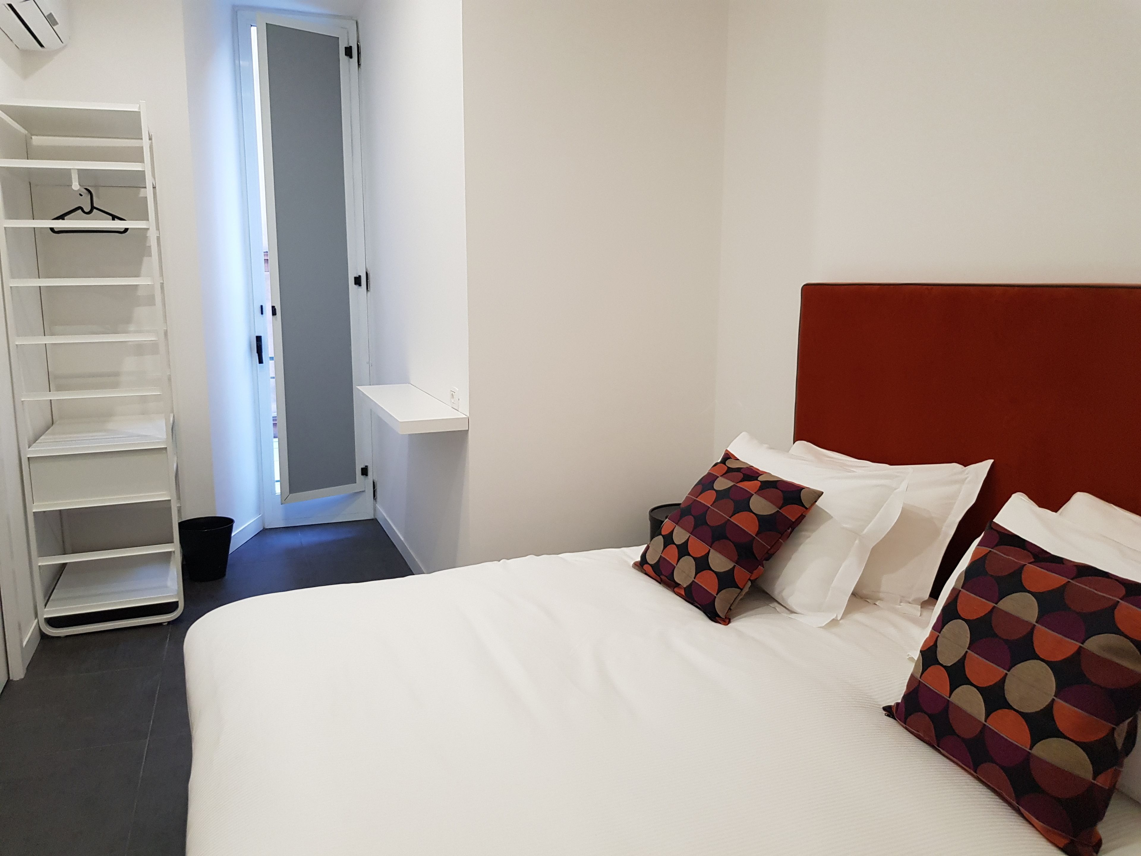A hotel room with a bed, two pillows, a white closet with shelves, and a door leading to another room.