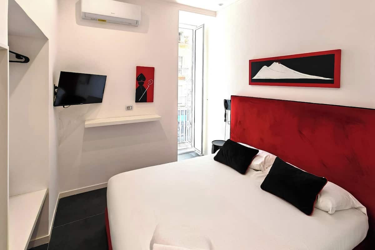 A modern hotel room with a large bed, a mounted TV, a red accent wall, and a view of the city through the window.