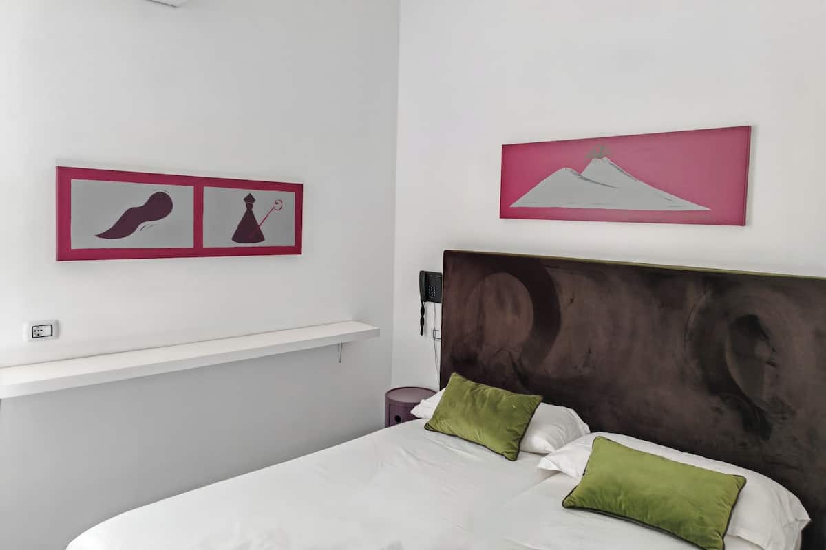 A bedroom with a bed, two pillows, a headboard, a bedside table, a wall-mounted phone, and two framed artworks.