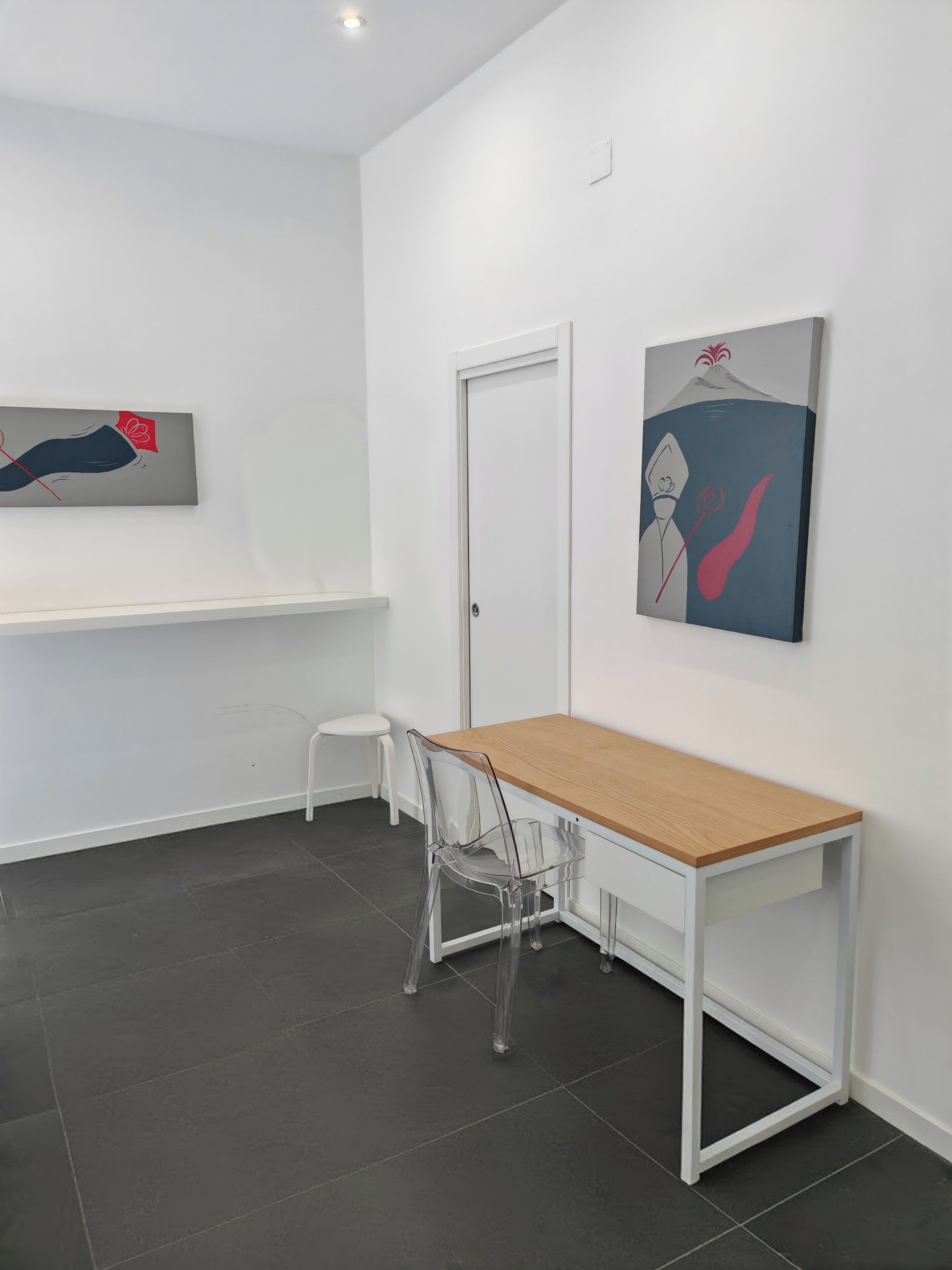 A minimalist room with a wooden table, a clear plastic chair, a white door, and two artworks on the walls.