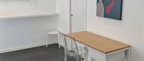 A minimalist room with a wooden table, a clear plastic chair, a white door, and two artworks on the walls.