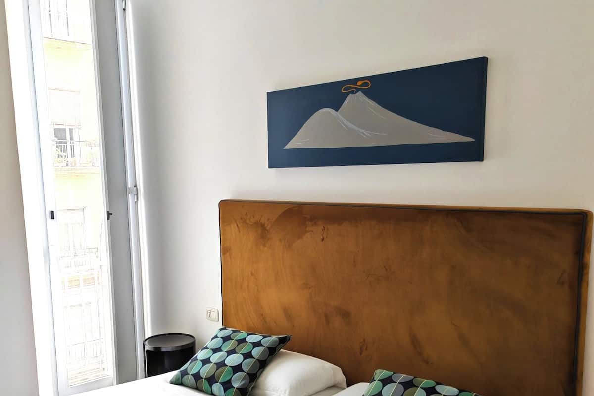 A hotel room with two beds, a wooden headboard, a painting of a mountain, and a window with a view of buildings.