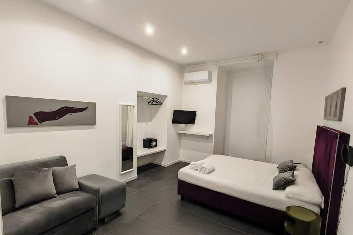 A modern, well-lit bedroom with a grey sofa, a bed with a white mattress, and a TV mounted on the wall.