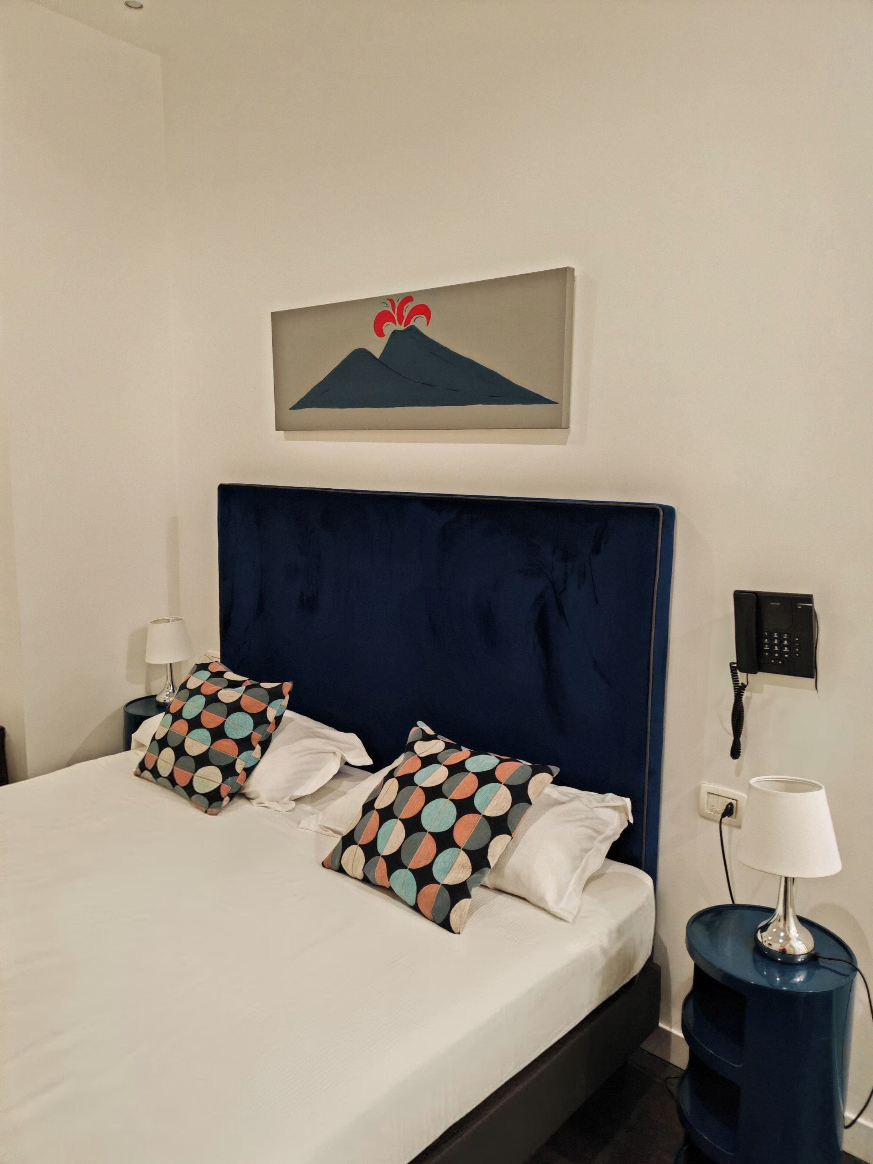 A neatly made bed with two pillows, a wall-mounted phone, and a lamp on a bedside table.