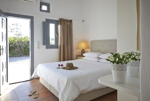 A hotel room with a bed, a nightstand with a lamp, a window with curtains, and a door leading to an outdoor area. - Aleria Villa (Santorini)