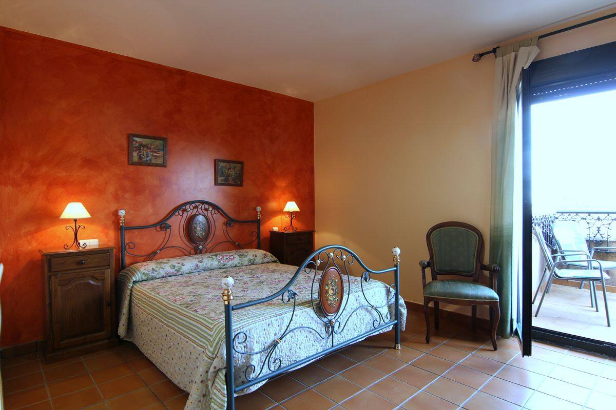 A bedroom with a bed, two bedside tables with lamps, a chair, and a balcony with a table and chairs.