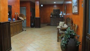 Un vestíbulo con mostrador de recepción, una zona de estar con una butaca y un jarrón grande, y un reloj en la pared.