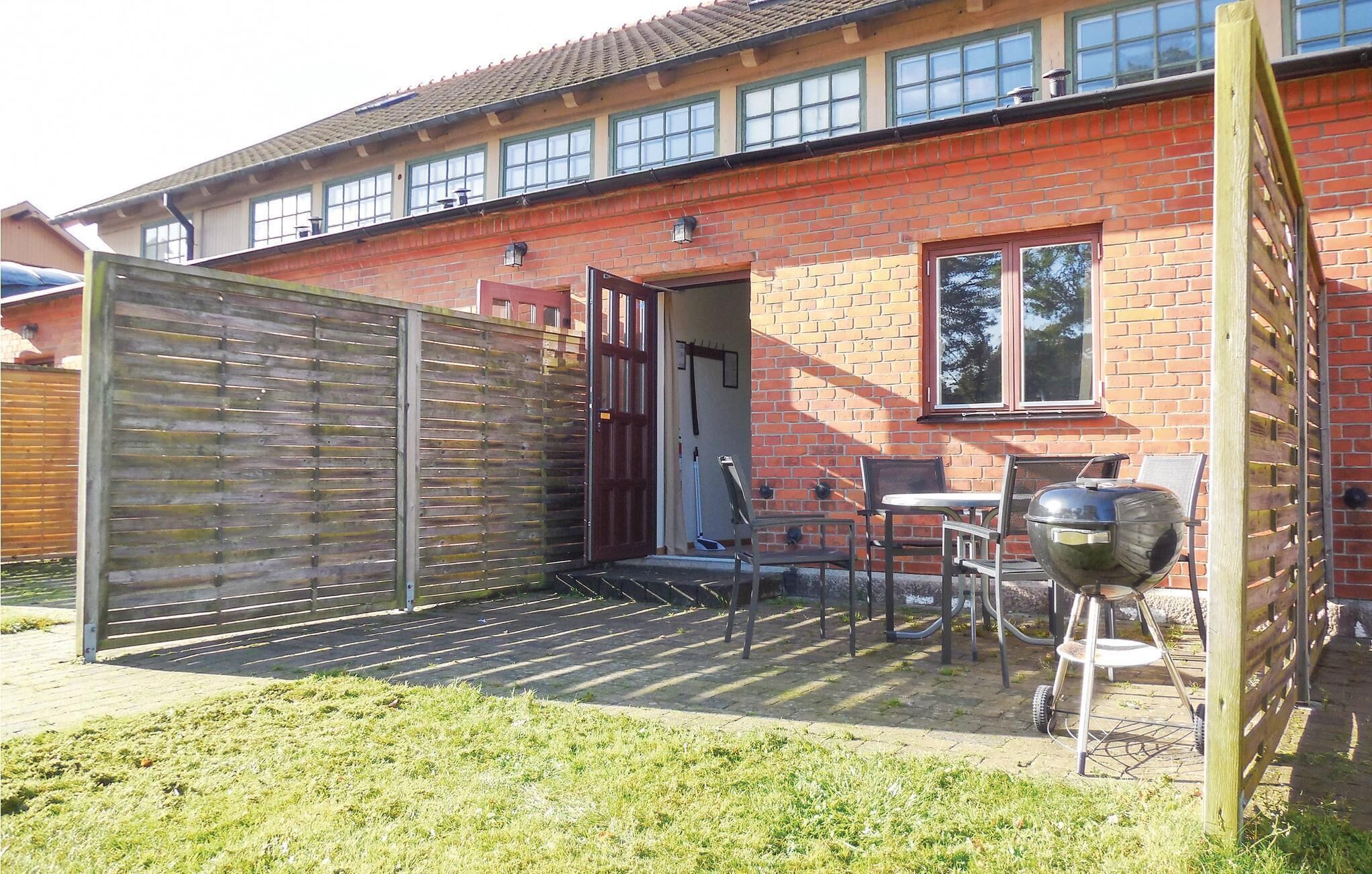 A brick house with a wooden gate, a patio with chairs and a barbecue grill, and a window with a view of trees.