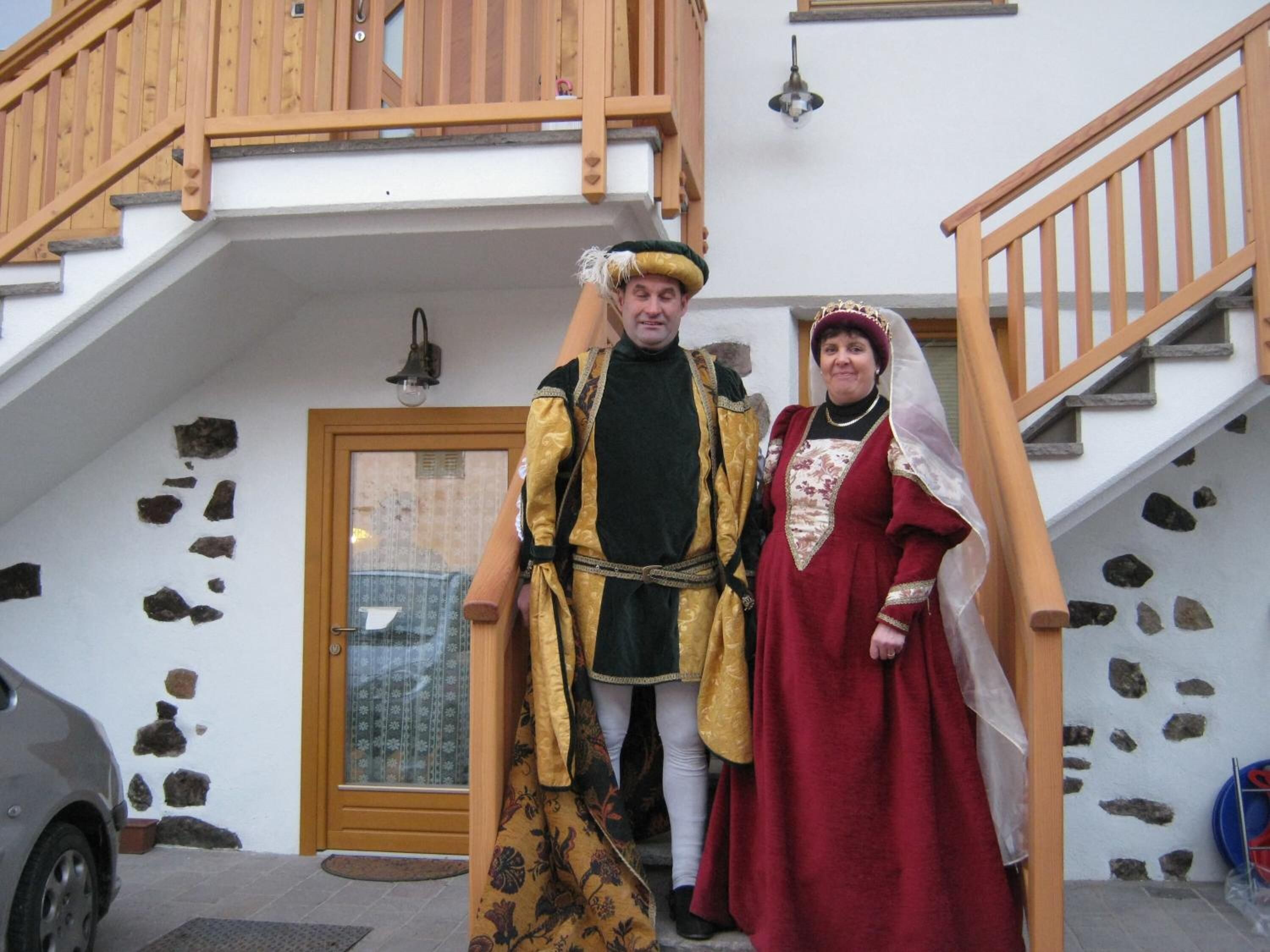 Un uomo e una donna in costumi d'epoca davanti a un edificio con una scala di legno e pareti in pietra.