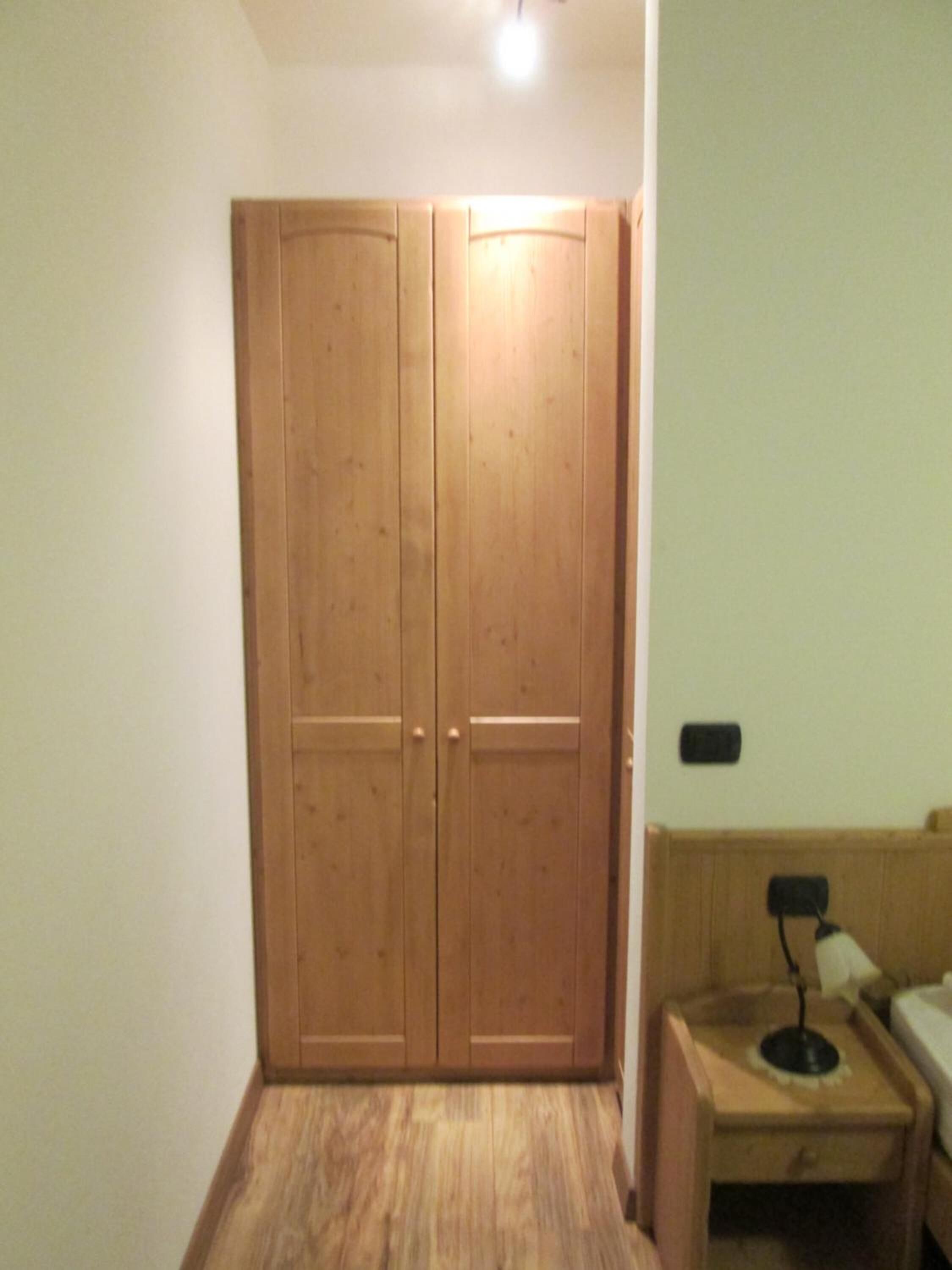 A room with a wooden wardrobe, a bedside table with a lamp, and a bed.