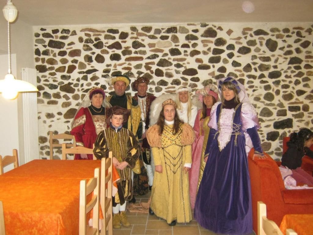 Un grupo de personas con trajes históricos en una habitación con pared de piedra y una cama con colcha naranja.