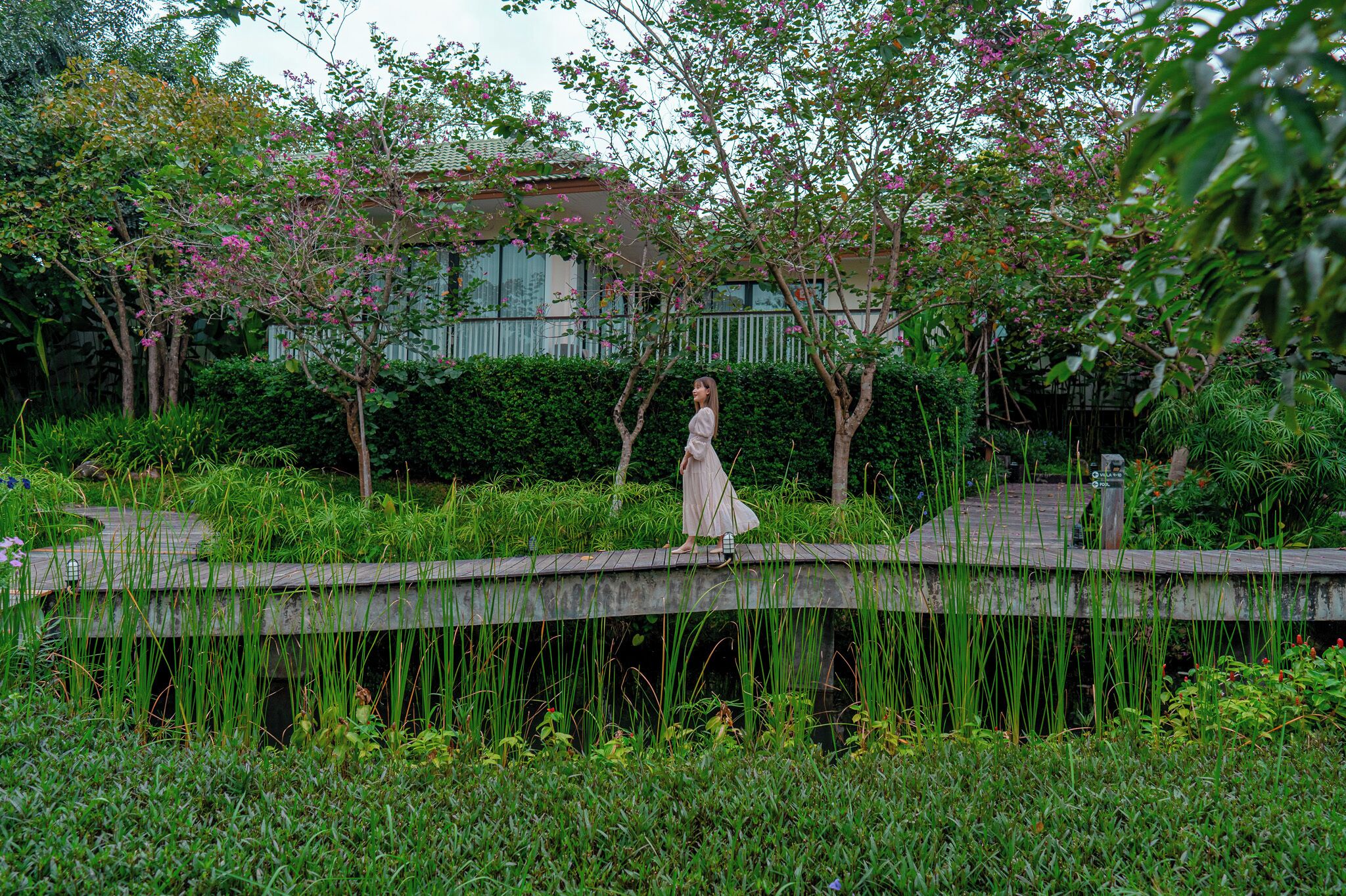 A woman standing on a wooden bridge in a lush garden with flowering trees and a building in the background.
