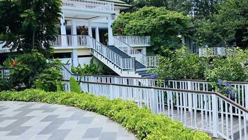 A multi-story hotel with a central staircase, surrounded by lush greenery and a paved walkway.