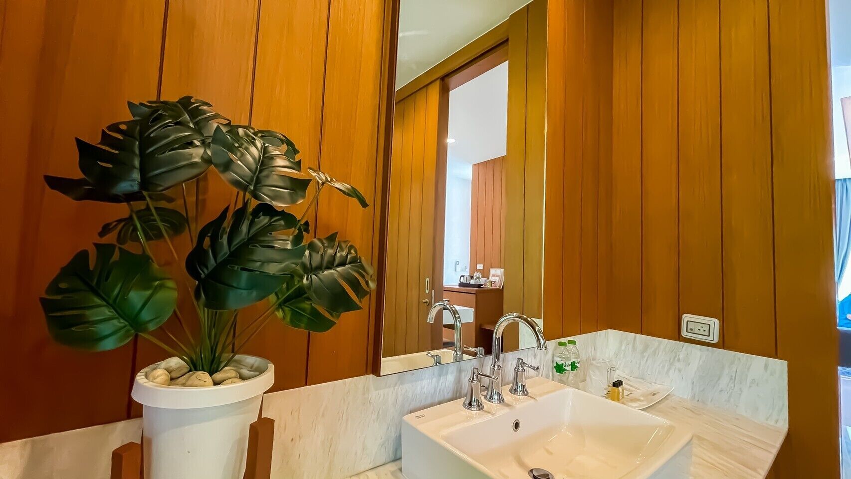 Un baño con un lavamanos blanco, dos grifos, un espejo grande, paneles de madera y una planta en maceta con hojas anchas.