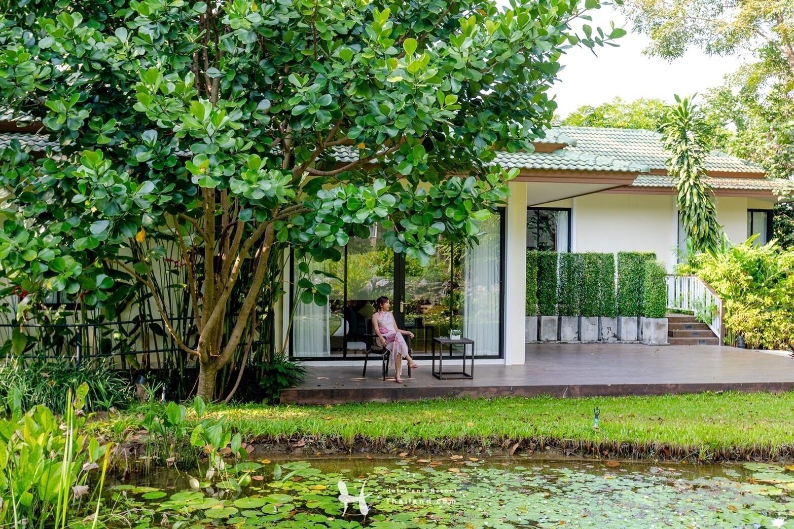 A serene garden with a pond, lush greenery, and a modern house with a covered patio.