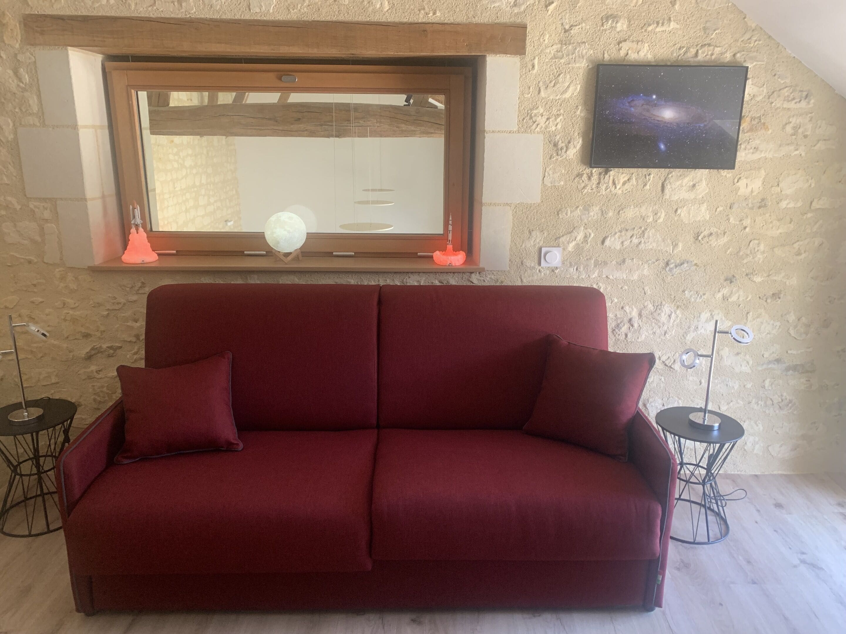 Un sofá rojo con dos almohadones, una ventana con marco de madera y vistas, un televisor fijado a la pared y dos mesitas con lámparas.