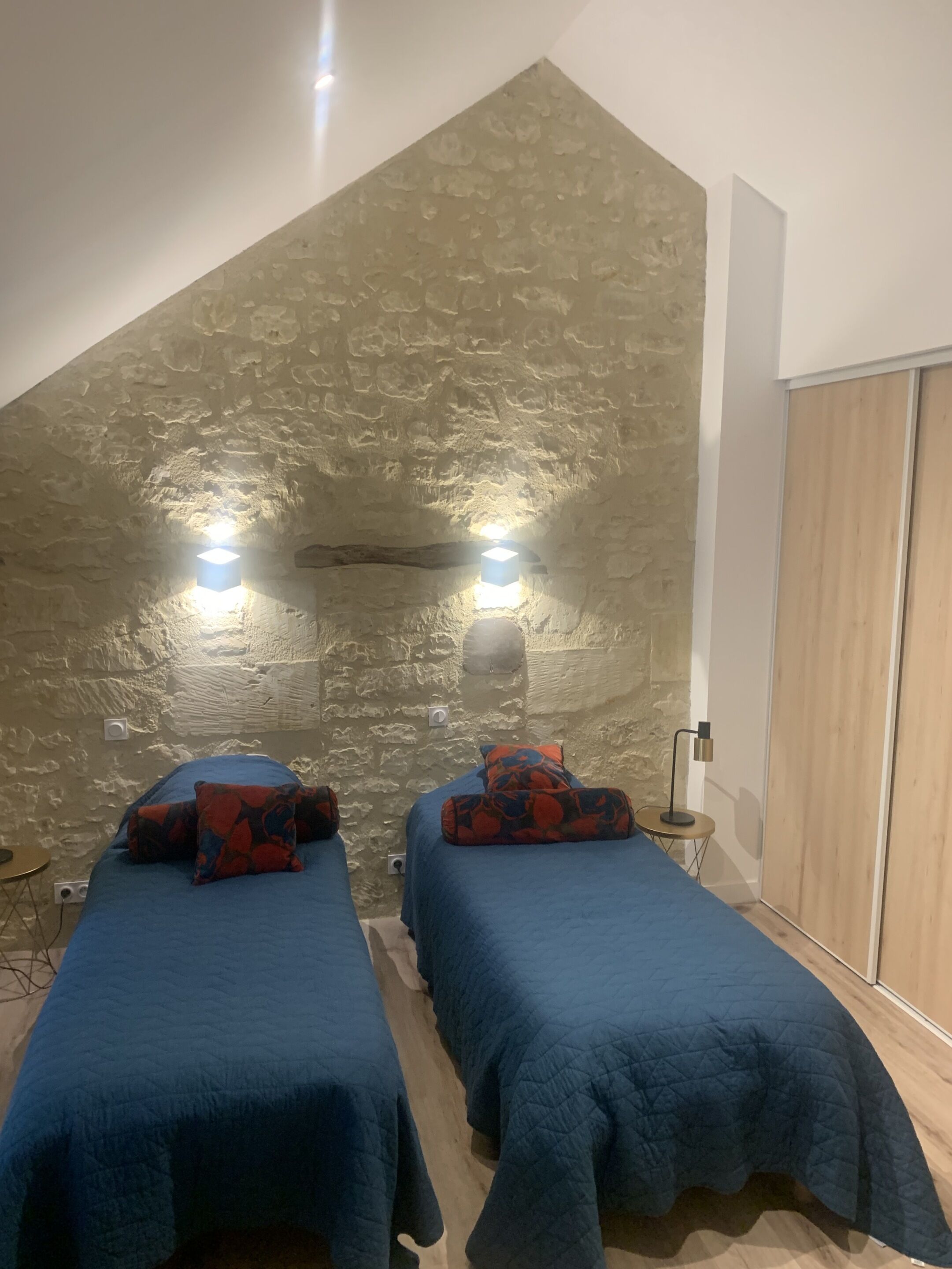 A room with two beds, each with a blue bedspread and red and blue patterned pillows. There are two wall-mounted lights and a small table with a lamp. The room has a stone wall and a wooden door.