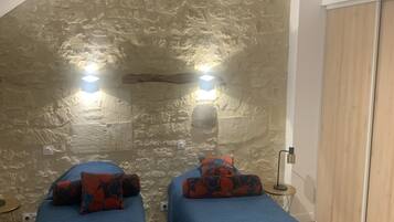 A room with two beds, each with a blue bedspread and red and blue patterned pillows. There are two wall-mounted lights and a small table with a lamp. The room has a stone wall and a wooden door.