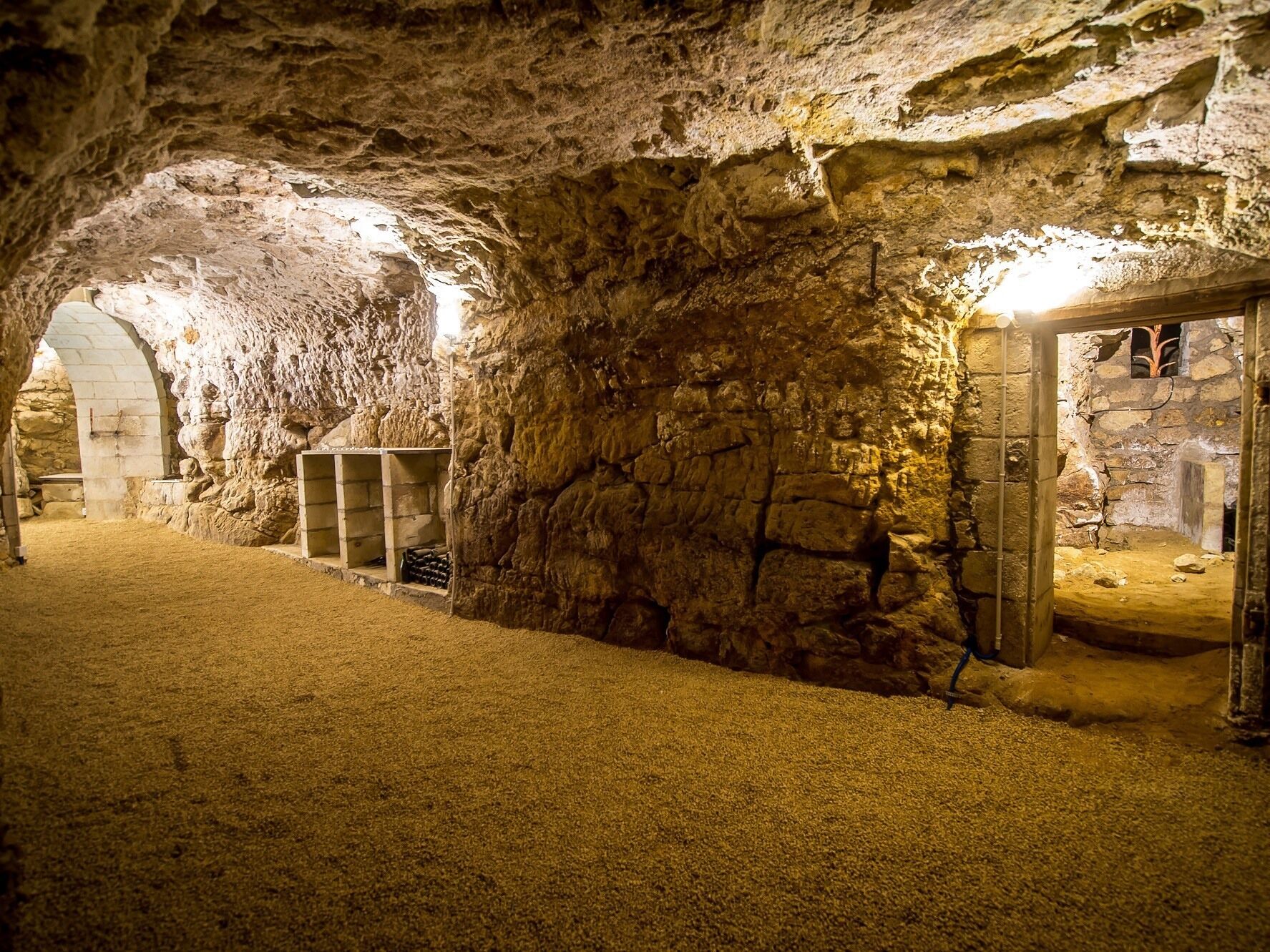 Un interior que recuerda a una cueva, con paredes de piedra, una puerta de madera y un estante con botellas.