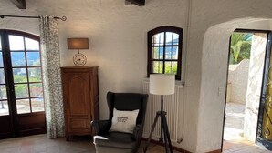Une pièce comprenant un meuble en bois, un fauteuil noir avec un coussin, un lampadaire et deux fenêtres donnant sur un paysage verdoyant.