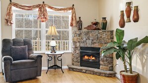 A cozy living room with a stone fireplace, a comfortable armchair, a table with a lamp, and a large plant.