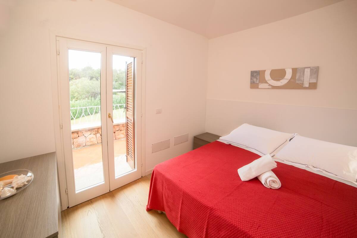 Camera da letto con copripiumino rosso, cuscino bianco e asciugamano sul letto. Una porta dà accesso a un'area esterna con vista sul verde.