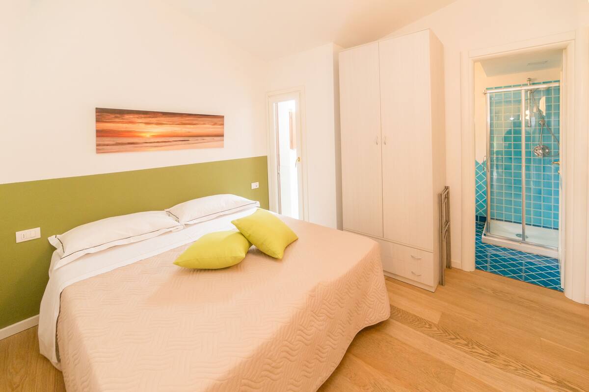Camera da letto con un letto, parete verde, armadio bianco e bagno con piastrelle blu.